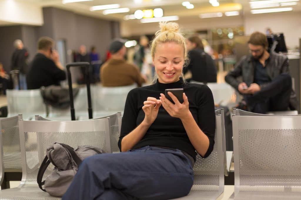 young white woman with blond hair looking at her phone in an airport, she is wearing a black sweater and jeans, download entertainment or apps for the flight ahead