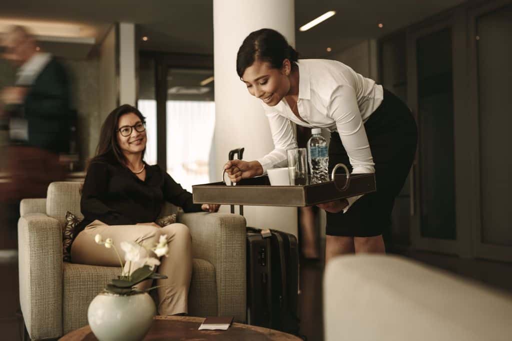 Two women in an airport lounge, one is sitting down on a nice comfortable chair and the other is a waitress bringing a tray with drinks; if you’re facing a long delay, one of the best ways to make the experience more comfortable is to escape the busy terminal and head to an airport lounge