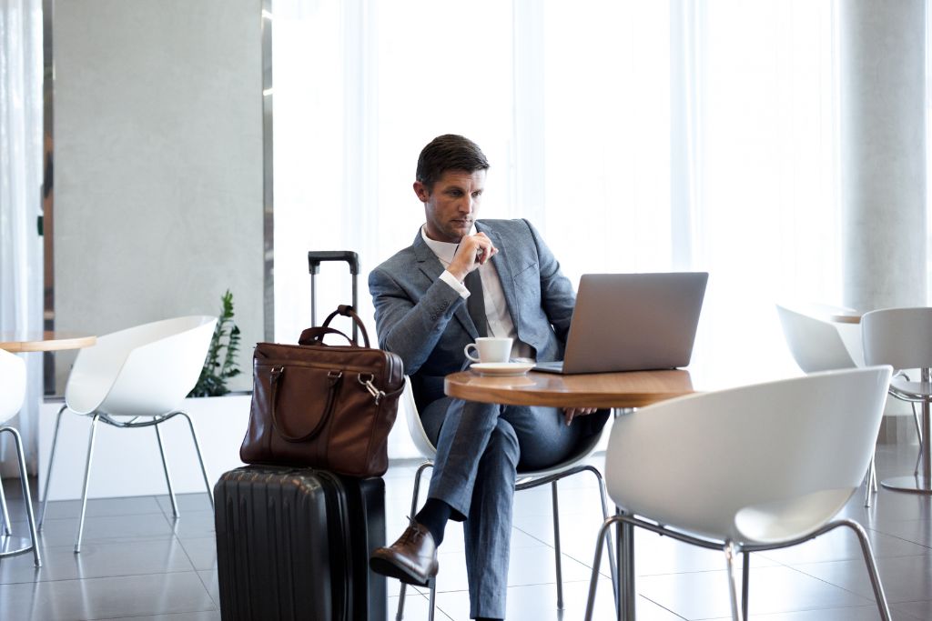 A white man sitting in an airport café working on his laptop, he is dressed in a gray suit and his luggage and bag are beside him; instead of wasting time, pull out your laptop or tablet and knock out some of those lingering tasks