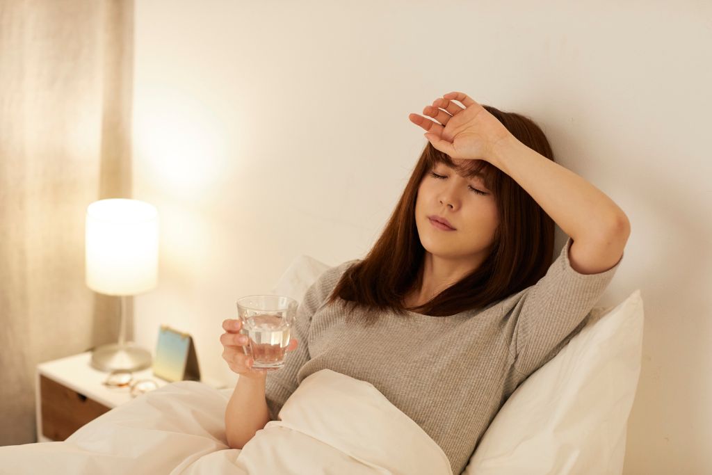 A young Asian woman laying in a bed feeling sick, holding a glass of water and touching her forehead for fever, even if you're healthy, bring common over-the-counter meds. It's much easier than trying to find a pharmacy abroad when you’re not feeling well