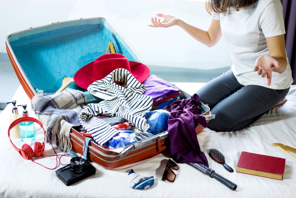 A woman sitting on a bed in front of a red luggage that has all sorts of items inside, overpacking