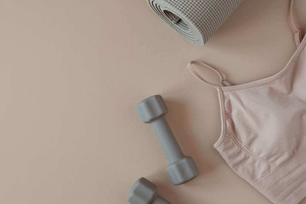 A close up shot of two dumbbells, a sports bra and the corner of a yoga mat, everything is in neutral colors, what not to bring is heavy workout gear