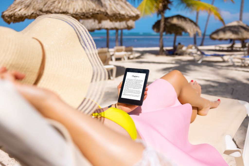 A woman with a sunhat, readying a kindle on a beach