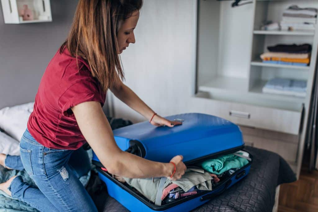 A white woman trying to close her blue luggage on a bed, what not to pack is too many clothes, overpacking