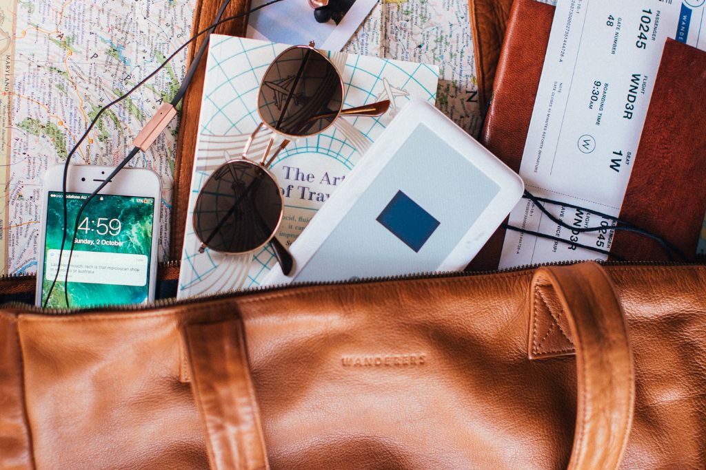 A close up of a personal item (a leather bag), inside there is a cell phone, sunglasses, a boarding pass, a book