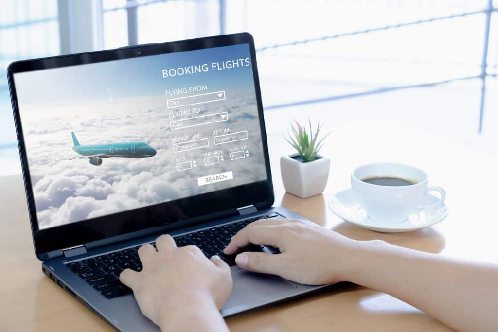 A person typing on a laptop, the screen shows a website that says flight booking, there is a cup of coffee beside the person