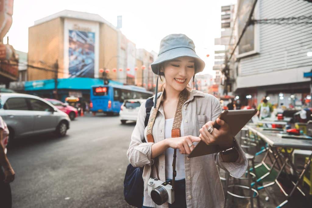 A young Asian woman with a camera and an iPad exploring a city, there are cars and traffic behind her