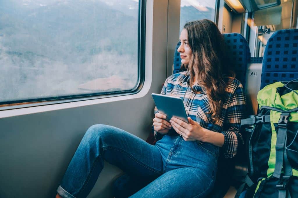 A solo female traveler in a train looking out of the window and holding a tablet, her backpack is beside her